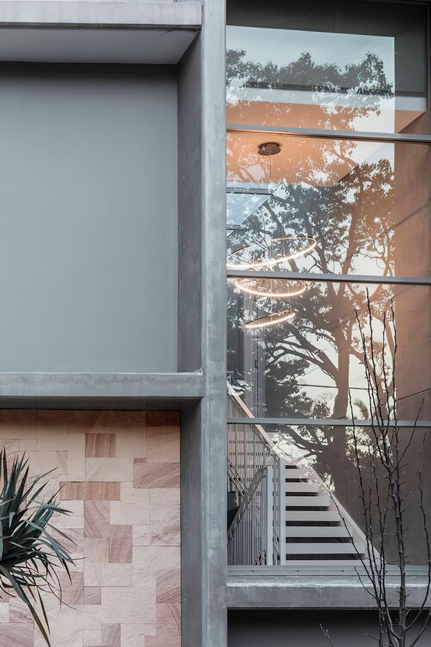 A close-up exterior architectural view of a contemporary concrete building with expansive glazed openings, an internal stairway, exposed structural framing, and a geometric feature wall clad in pale timber-toned tiles. Reflections of trees and circular pendant lights are visible in the glazing, with greenery partially framing the facade.