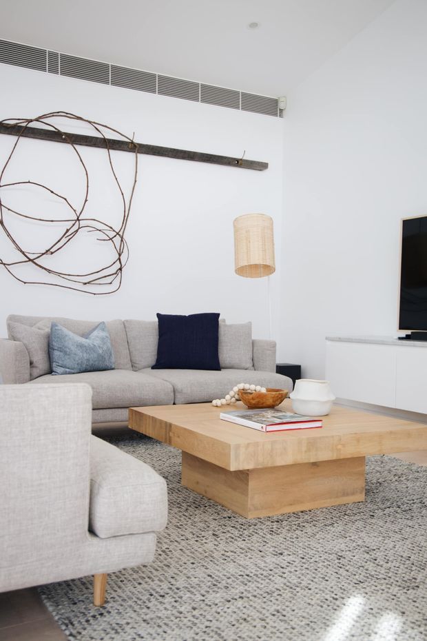 Bright contemporary living room with a large light-grey sectional sofa, navy and pale blue cushions, a substantial natural timber coffee table, and a textured grey area rug. White walls feature an exposed dark timber rail with sculptural branch-like artwork, while a woven pendant light and minimalist white media console complete the room.