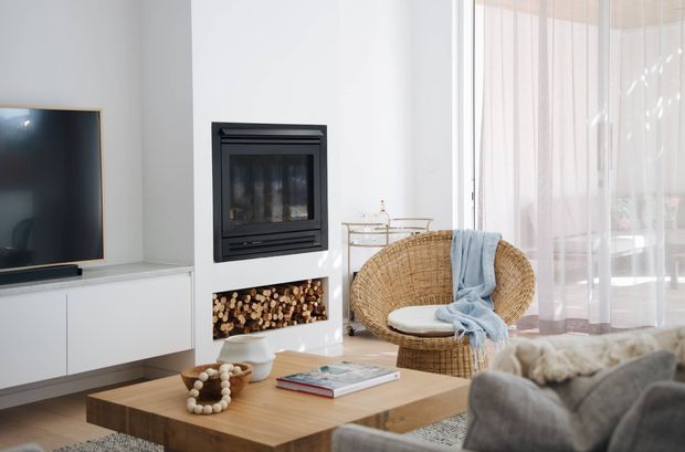 Bright contemporary living room featuring a wall-mounted television on white built-in cabinetry, a black inset fireplace with stacked firewood storage below, a woven rattan lounge chair, light timber coffee table, and sheer floor-to-ceiling curtains. The space has a warm, relaxed modern feel with abundant natural light.