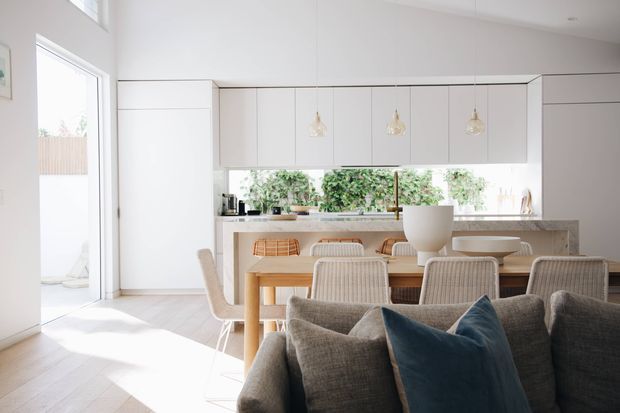 Bright open-plan interior showing a contemporary white kitchen with full-height cabinetry, a stone island or benchtop, pendant lights, and a timber dining table. A grey upholstered sofa with cushions occupies the foreground, while large glazing and greenery beyond the kitchen create an airy connection to the outdoors.