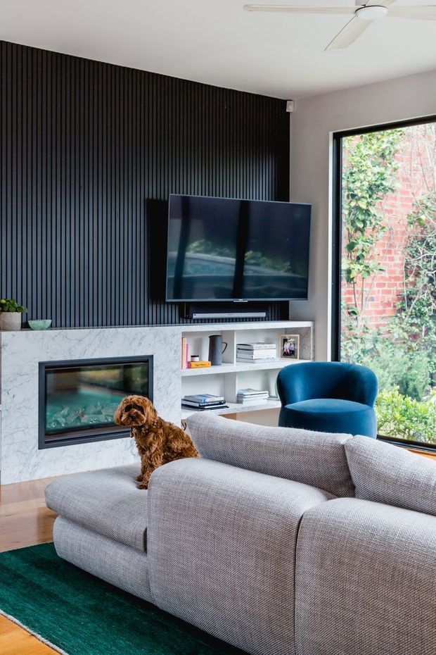 A contemporary living room with a large grey sectional sofa, teal area rug, deep blue upholstered armchair, and a wall-mounted television. The feature wall combines dark vertical timber slats with a white marble-look fireplace surround and built-in open shelving. Large black-framed glazing overlooks a leafy garden and brick wall, while a ceiling fan and small dog complete the scene.