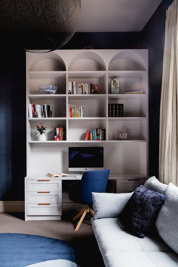 A refined home office integrated into a living room, featuring a white built-in shelving and desk unit with arched display niches, books, drawers, and a desktop computer. The room has deep navy walls, a large textured pendant light, full-height cream curtains, a blue upholstered desk chair, and a pale grey sectional sofa with dark cushions. A blue area rug and warm timber accents complete the sophisticated, moody interior.