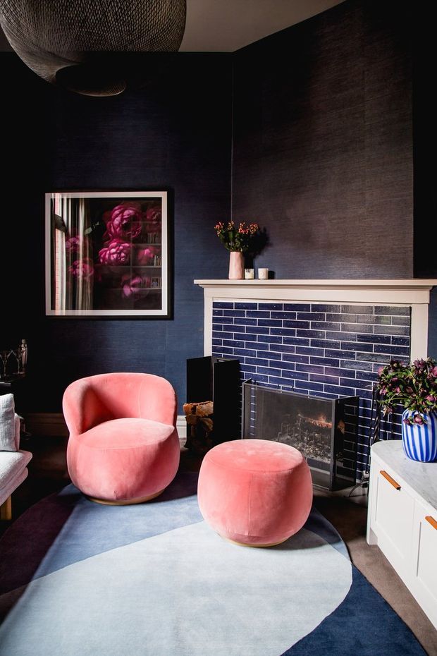 A richly styled living room with deep navy textured walls, a dark blue glazed brick fireplace surround, and a pale mantel. Two rounded blush-pink lounge chairs and a matching ottoman sit on a large abstract blue and light-grey rug. A framed floral artwork, sculptural pendant light, plants, and white built-in cabinetry complete the dramatic, colourful interior.