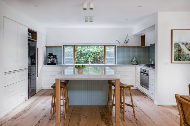 Bright contemporary kitchen with white full-height cabinetry, a large central island with a pale stone benchtop and vertical panelled base, integrated appliances, and a muted green square-tile splashback. Timber bar stools and wide-plank timber flooring add warmth, while a timber-framed window brings in garden views.