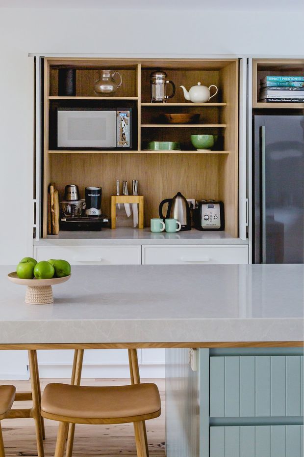 A bright contemporary kitchen with a built-in open timber display cabinet containing a microwave, coffee machine, kettle, toaster, crockery and glassware. White lower cabinetry and a pale stone island benchtop contrast with the warm wood shelving and timber flooring. A bowl of green apples and tan upholstered dining stools sit in the foreground, while a muted blue-grey island panel adds colour.