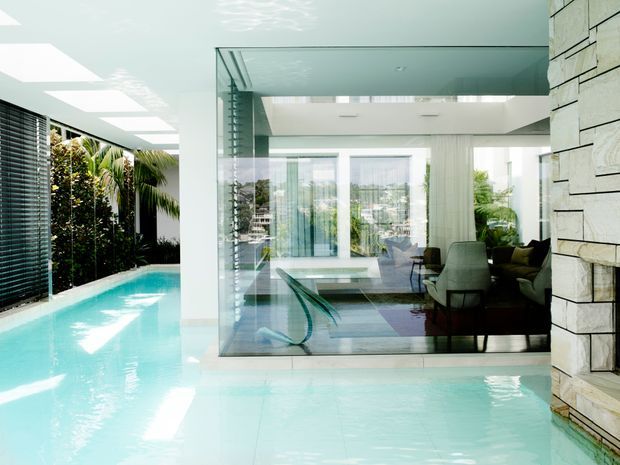 Bright contemporary interior with an indoor lap pool running alongside a glazed living room. The space features floor-to-ceiling glass walls, a pale tiled pool surround, white ceilings with multiple skylights, a dark louvred screen, lush planting visible along the pool edge, and a substantial light stone-clad wall with horizontal joints.