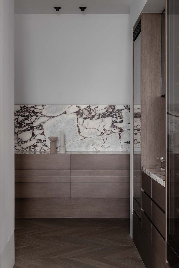 A refined compact bathroom or dressing vanity area viewed through a doorway, featuring dark timber-look cabinetry with integrated drawers, a dramatic white marble backsplash with burgundy-grey veining, a matching stone vanity benchtop, dark herringbone flooring, and a large mirrored cabinet. Minimal black ceiling spotlights and neutral plaster walls complete the moody, luxurious scheme.