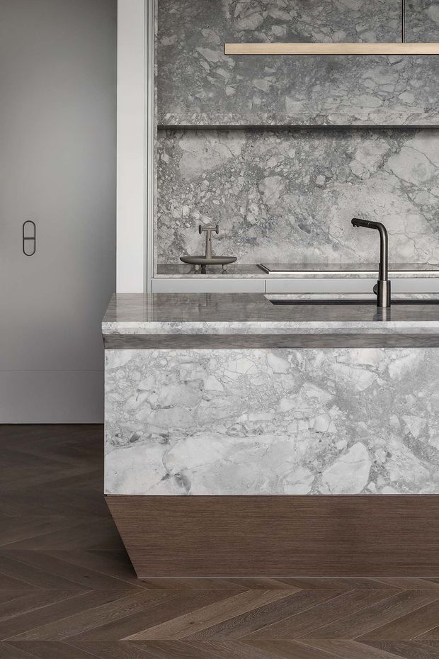 Close-up view of a sophisticated contemporary kitchen island featuring a heavily veined grey natural-stone benchtop and waterfall-style stone cladding over a dark timber base. Matching stone forms the splashback and open shelving backdrop, with a dark metal tap, integrated sink and understated linear pendant light. Dark timber herringbone flooring and muted grey cabinetry create a refined, tonal interior.