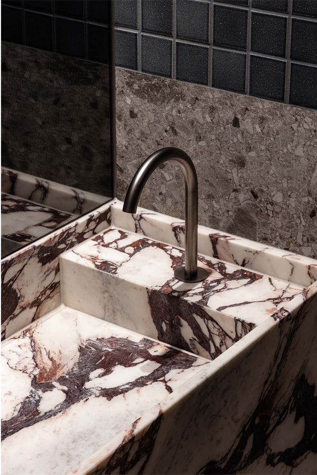 Close-up of a sculptural bathroom vanity and integrated basin clad in dramatic veined natural stone, paired with a dark brushed-metal arched tap. The wall features dark square tiles above a matching stone backsplash, with a mirror visible at left.