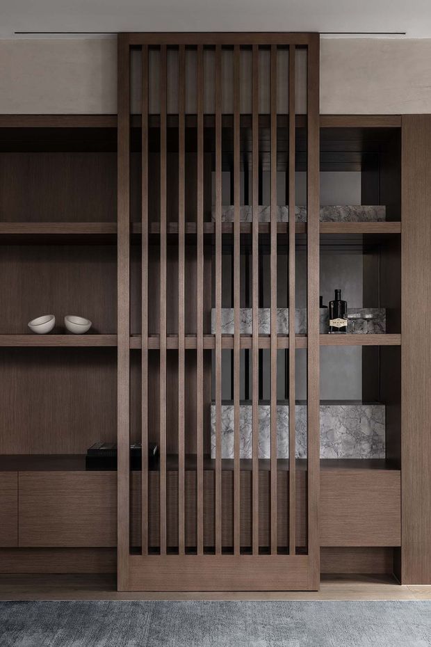 A refined built-in storage and display wall in a dark, warm-toned wood finish. Open shelving on both sides contains minimal decorative objects and bottles, while a central full-height timber slatted screen creates visual separation and depth. Marble-look stone shelves or backing provide contrast, with a neutral plaster wall and grey carpet completing the understated interior.