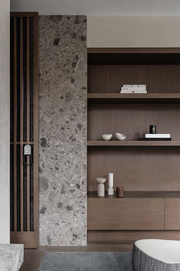Close-up of a refined contemporary interior featuring a full-height grey terrazzo or aggregate-stone wall panel beside a dark timber slatted door. Built-in dark wood shelving and cabinetry occupy the right side, styled with minimalist ceramics and books. A stone-topped surface, muted rug, and upholstered seating edge appear in the foreground, creating a warm, textural, understated composition.