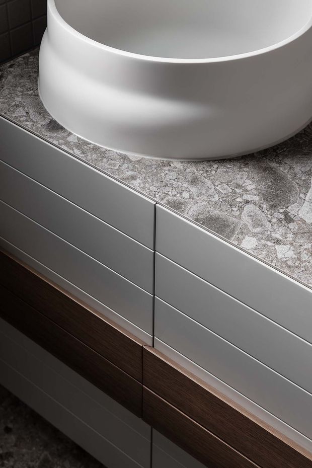 Close-up detail of a contemporary bathroom vanity with a white vessel basin, grey stone-patterned benchtop, smooth grey drawer fronts, and dark timber cabinetry below. A dark tiled wall and matching floor are partially visible.