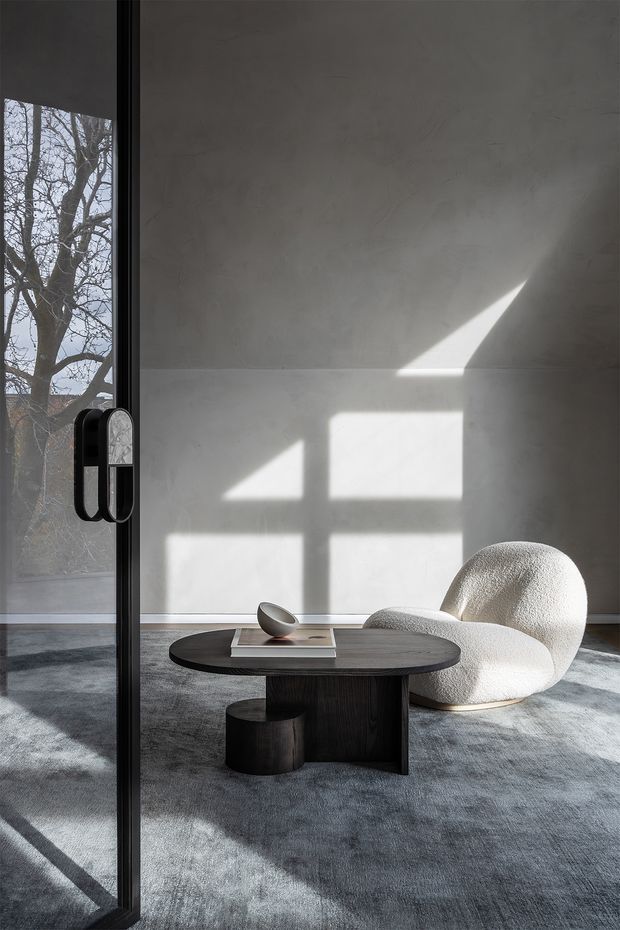 A minimalist contemporary interior lounge viewed through a black-framed glazed door. The room features a pale textured plaster wall, expansive grey carpet, a low dark timber coffee table with cylindrical pedestal supports, and a sculptural off-white boucle lounge chair. Strong sunlight casts geometric window shadows across the wall and floor, creating a calm, gallery-like atmosphere.