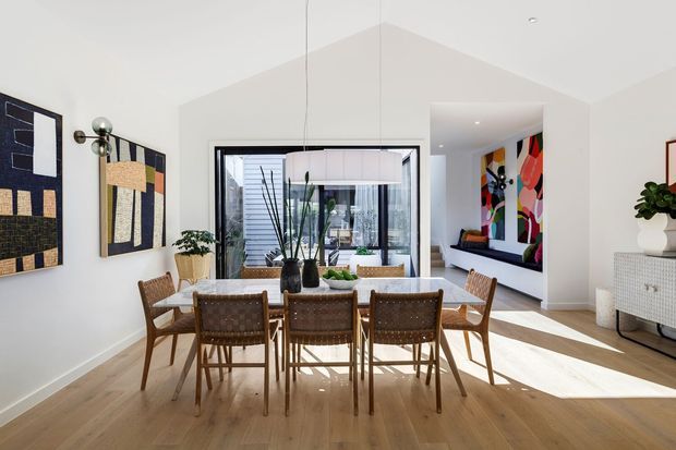 Bright contemporary dining area with a long timber dining table and woven timber chairs, set on a light timber floor. White walls and a high pitched ceiling create an airy volume, while abstract artwork, indoor plants, and a large glazed opening add visual interest. A living area with colourful artwork is visible beyond the dining space.
