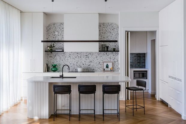 Bright contemporary kitchen with a large white island, vertical fluted detailing, four dark bar stools, terrazzo-look splashback and shelving, white cabinetry, and warm timber parquet flooring. A doorway leads to a darker tiled utility or pantry area.