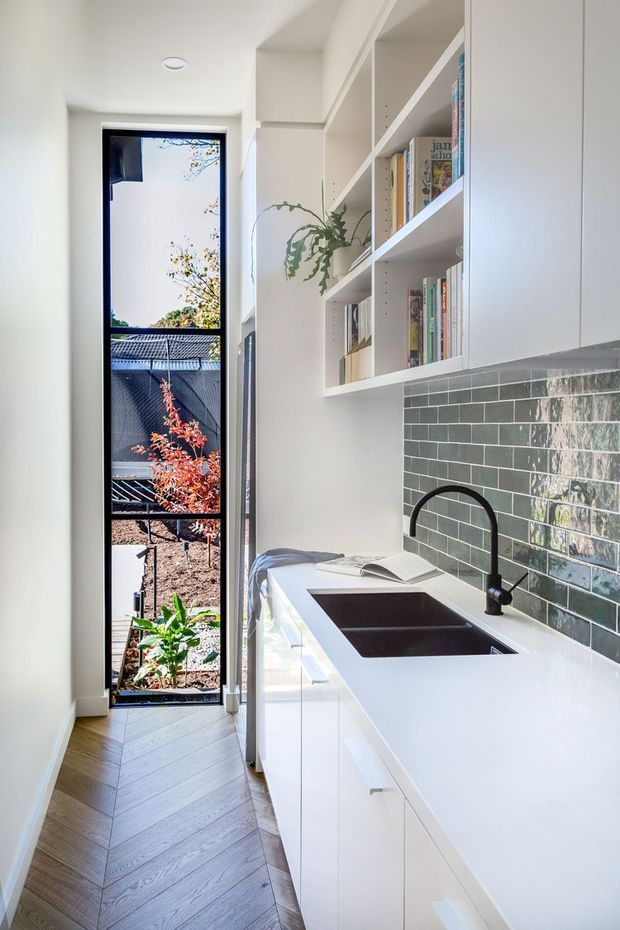 Bright, narrow contemporary kitchen with a long white stone-look benchtop, undermount double sink, matte black gooseneck tap, white handleless cabinetry, and open white shelving displaying books and a trailing plant. A glossy dark green subway-tile splashback runs along the work area. The room has herringbone timber flooring and a tall glazed door or window opening to a small planted outdoor area.