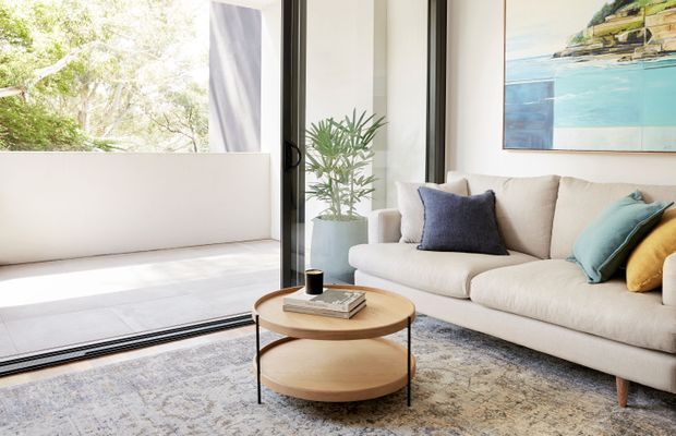 Bright contemporary living room with a large cream upholstered sofa, colourful accent cushions, a round two-tier timber coffee table, and a textured neutral rug. Full-height black-framed glazing opens to a light-filled outdoor terrace, while a potted plant and coastal artwork add natural and blue-green accents.