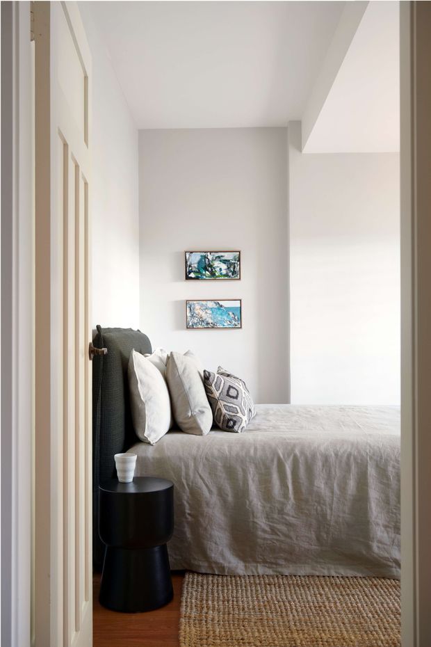 A calm, light-filled bedroom viewed through a doorway. The room features a neatly made bed with a textured light grey-beige cover, layered white and patterned cushions, a dark upholstered headboard, a small black cylindrical bedside table, timber flooring, and a natural woven rug. Two small framed abstract artworks hang on the pale wall, with subtle vertical wall panelling visible along the left side.