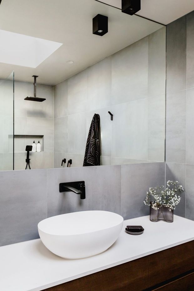 Contemporary bathroom vanity with a white vessel basin set on a white countertop above dark timber cabinetry. Large pale grey tiled walls, black wall-mounted tapware and shower fittings, a frameless glass shower screen, recessed niche, skylight, and black ceiling spotlights create a clean, refined look.