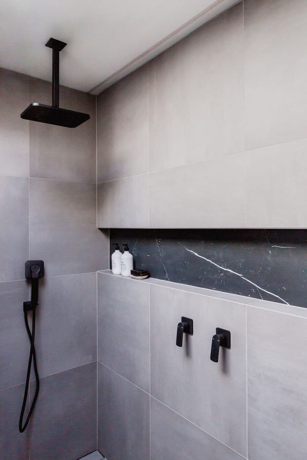 Close-up view of a modern walk-in shower with large light-grey wall tiles, matte black ceiling-mounted rainfall showerhead, matching handheld shower, and black shower controls. A horizontal dark marble-look recessed shelf with white veining provides a contrasting feature surface and storage niche.