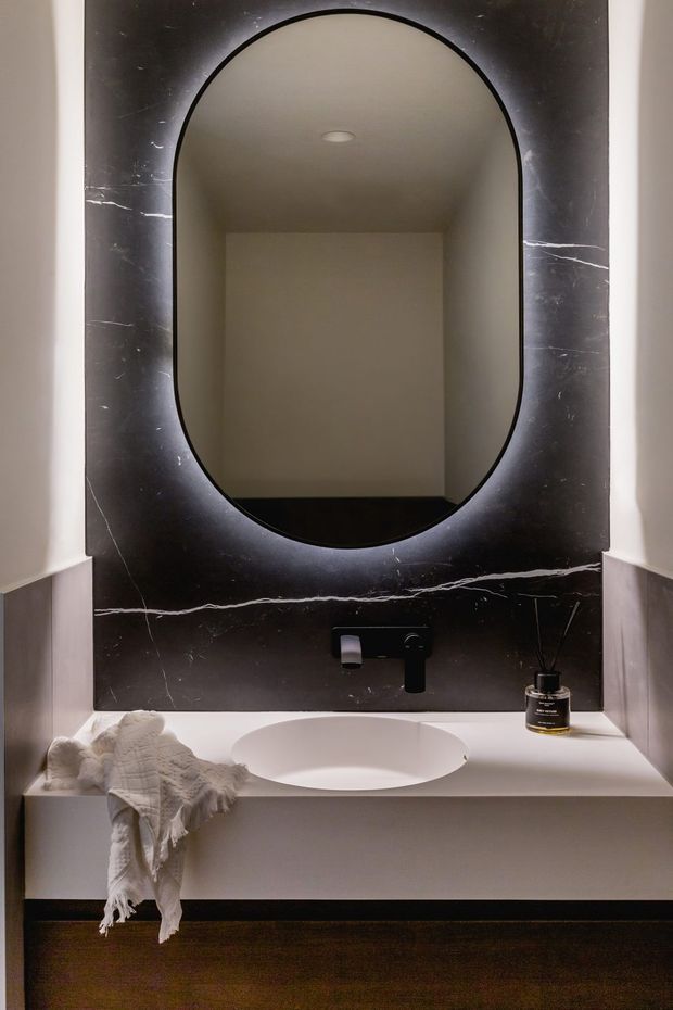 A compact, refined bathroom vanity area featuring a white integrated basin and countertop, matte black wall-mounted tapware, and a large rounded-arch mirror set into a dramatic dark marble-look slab with white veining. Soft neutral walls, warm timber flooring, recessed ceiling lighting, and folded towels create a calm, luxurious atmosphere.