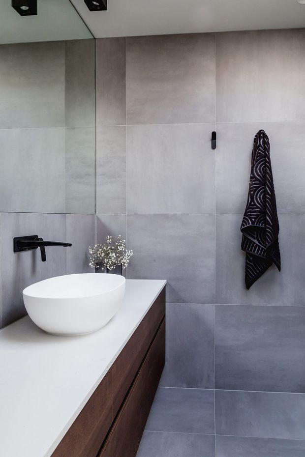 A contemporary bathroom featuring large grey wall and floor tiles, a white vessel basin on a white benchtop, dark timber vanity cabinetry, matte black wall-mounted tapware, a large mirror, and a patterned dark towel. The clean-lined fit-out has a refined monochromatic palette with warm timber contrast.