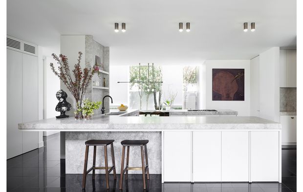 Bright contemporary kitchen with a large white stone island and integrated cabinetry, two dark timber bar stools, black tiled flooring, white walls, and extensive glazing overlooking greenery. A sculptural pendant, artwork, decorative branches, and open shelving add restrained visual accents.