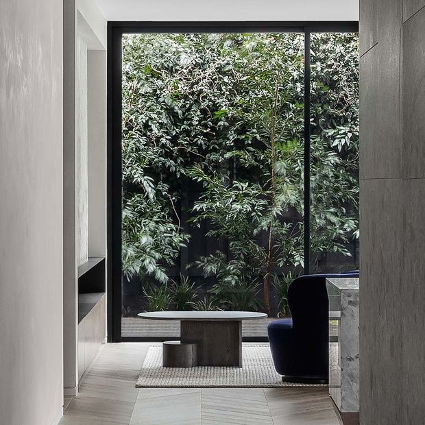 A refined contemporary interior vignette facing a lush private garden through full-height black-framed glazing. The space features pale stone or timber-look flooring, a textured neutral rug, a sculptural dark upholstered lounge chair, a low wood-toned table, built-in shelving, and a pale stone-clad counter or wall edge. The restrained palette and abundant greenery create a calm, sophisticated atmosphere.