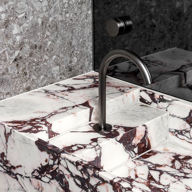 Close-up architectural detail of a sculptural bathroom vanity carved from heavily veined white stone, with an integrated stepped basin and a dark brushed-metal wall-mounted tap. A dark reflective wall panel and textured grey stone wall form the backdrop.