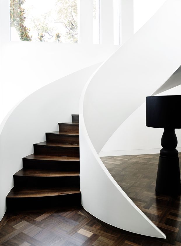 A sculptural curved staircase with dark timber treads rises beside a smooth white sweeping balustrade or curved wall. The space features dark parquet flooring, tall white walls, narrow vertical windows, and a black floor lamp visible beyond the stair. The composition is highly minimalist and architectural, with strong flowing curves and contrasting black, white, and timber tones.