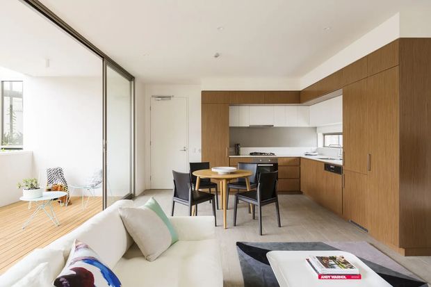 Bright open-plan living and dining area with a contemporary galley-style kitchen. A white sofa faces a low coffee table, while a compact round dining table with four dark chairs sits beside extensive warm timber cabinetry. Full-height glass sliding doors open to a light-filled balcony or sunroom with timber decking and minimal furniture. The space has recessed ceiling lighting, pale flooring, white walls, and clean-lined built-in joinery.