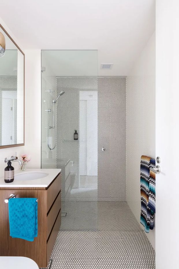 Bright contemporary bathroom featuring a walk-in shower enclosed by clear glass, with small circular mosaic tiles covering the shower walls and floor. A floating dark wood vanity with a white integrated basin sits to the left beneath a large framed mirror. Chrome shower fittings, white walls, a ceiling exhaust vent, and colourful patterned towels complete the space.