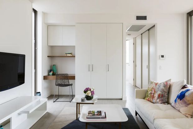 Bright, compact contemporary living room with a white built-in media console and wall-mounted television, a cream sofa with colourful patterned cushions, and a low white coffee table. The room also includes integrated floor-to-ceiling storage, a small built-in desk with shelving, pale flooring, and a hallway visible beyond.