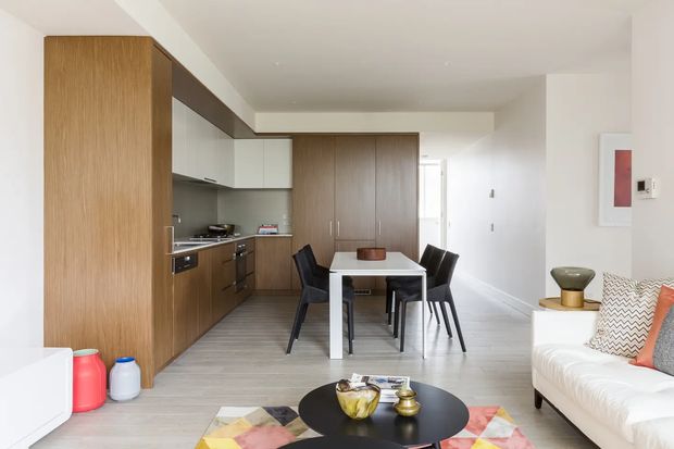 Open-plan contemporary apartment interior showing a compact kitchen with full-height timber cabinetry, a small dining table with four chairs, and an adjoining living area with a white sofa and round black coffee table. The space has light flooring, white walls, restrained furnishings, and a clean modern fit-out.