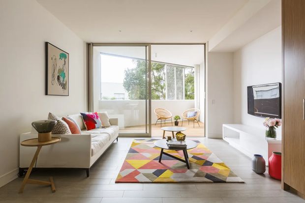 Bright contemporary living room with a white sofa, low black coffee table, colourful geometric rug, white built-in media cabinetry, and timber-toned storage cabinetry. Full-height glazed sliding doors open to a small balcony or terrace with additional seating, bringing in abundant natural light. The room has pale walls, a light timber-look floor, and restrained modern detailing with colourful accents.
