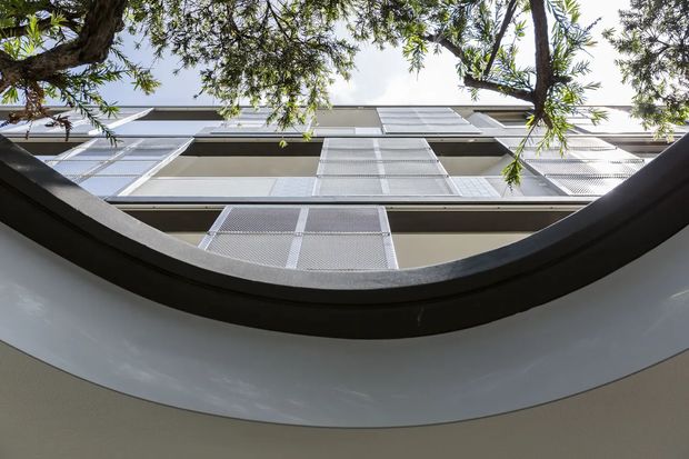 Upward-looking architectural view framed by a curved dark-edged opening, showing a multi-storey contemporary facade with repetitive balconies or openings, light grey metal mesh screens, and horizontal roofline elements. Tree branches and foliage extend across the upper edge against the sky.