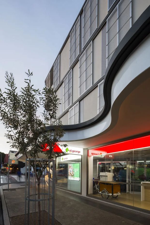 Street-level exterior view of a contemporary multi-storey commercial building with a curved projecting canopy, dark trim, pale facade panels, and extensive upper-level screening or louvred glazing. Ground-floor retail storefronts with prominent red illuminated signage open onto a paved urban pedestrian frontage with young street trees.