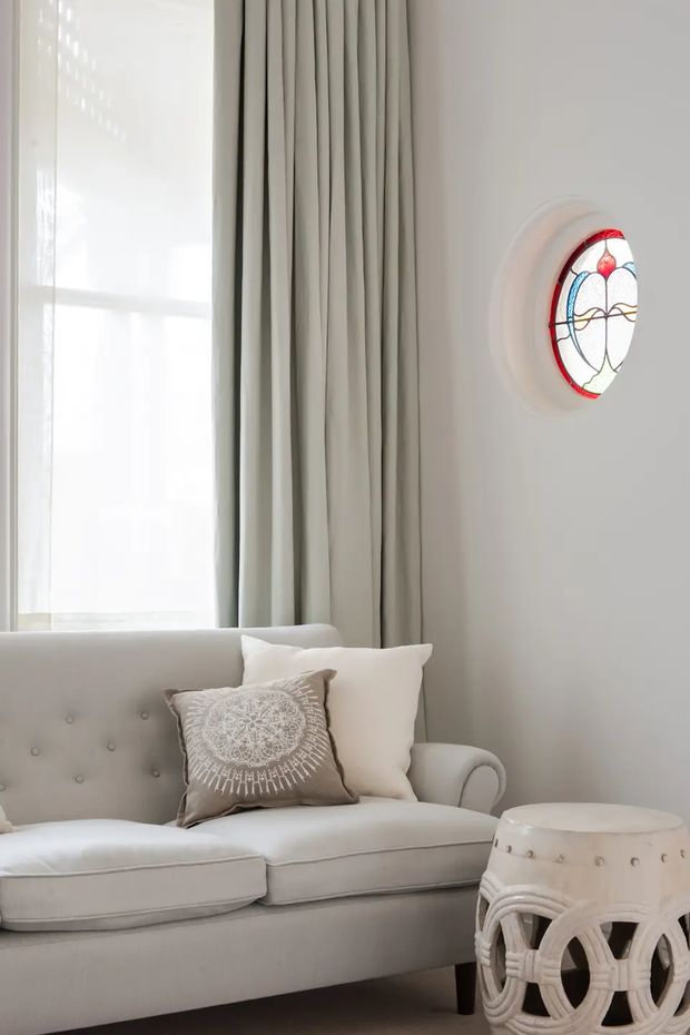 A bright, softly styled living room corner featuring a pale upholstered button-tufted sofa, layered neutral cushions, full-length pleated curtains, a large bright window, and an ornate white side table. A small circular stained-glass window adds a restrained traditional decorative detail to the otherwise light, calm interior.
