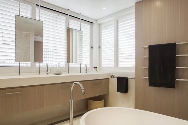 Bright contemporary bathroom with a long double vanity, wall-mounted rectangular mirrors, pale timber cabinetry, white benchtop, chrome tapware, extensive white plantation shutters, and a partially visible freestanding bathtub. A tall built-in timber storage and heated towel rail occupy the right side of the room.