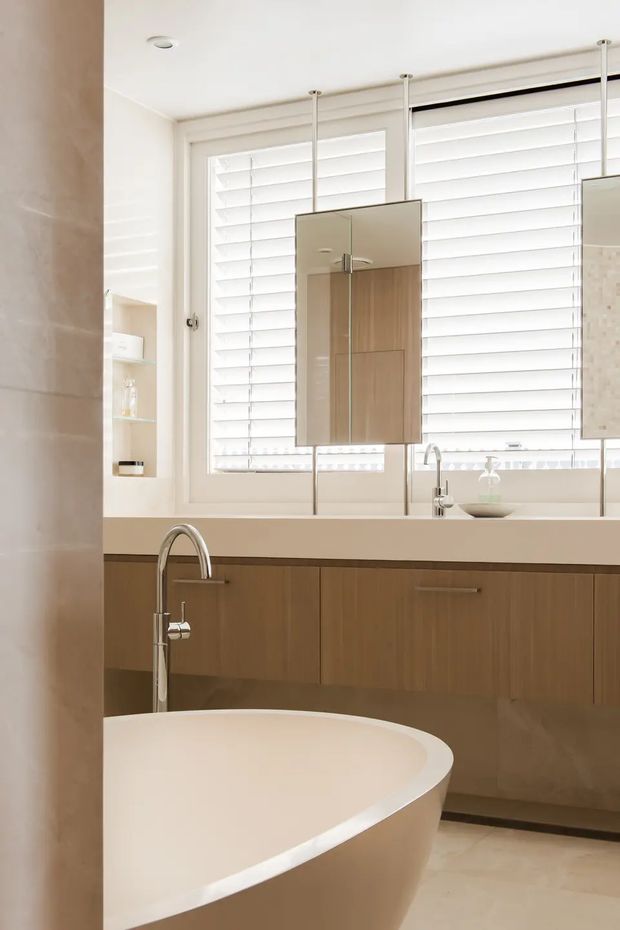 Bright contemporary bathroom featuring a freestanding oval bathtub in the foreground, a long timber-veneer vanity with a pale solid benchtop, integrated basin and chrome tapware, mirrored wall cabinets, and large windows fitted with white plantation shutters. The room has a clean, warm-neutral palette and abundant natural light.