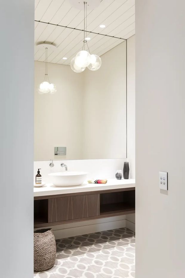 Bright contemporary bathroom vanity viewed through a doorway, featuring a large wall-to-wall mirror, white vessel basin on a white countertop, wall-mounted chrome tapware, dark timber floating cabinetry, patterned grey-and-white floor tiles, and a cluster of spherical pendant lights reflected in the mirror. The space has a clean, refined and softly illuminated appearance.