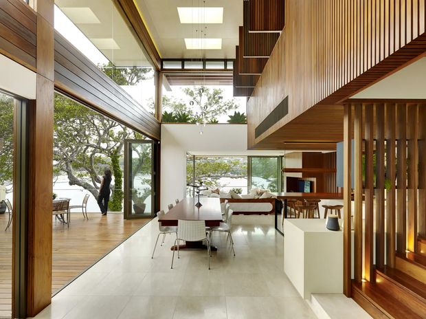 A sophisticated open-plan living and dining interior with a strong connection to the outdoors. The space features expansive floor-to-ceiling glazing, warm timber flooring and wall panelling, white architectural surfaces, a dining table with modern chairs, and a lounge area beneath a dramatic timber-lined upper volume. Vertical timber screens and built-in shelving define the circulation and stair area, while abundant daylight creates an airy, refined atmosphere.