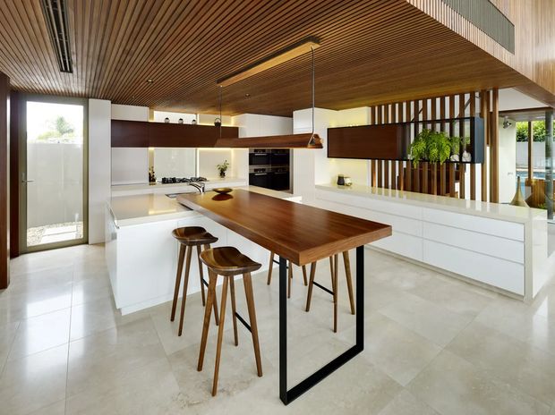 Contemporary open-plan kitchen with a large white island, integrated appliances, and a prominent timber dining table extension supported by a black metal frame. The space features a warm slatted timber ceiling, white cabinetry, pale tiled flooring, timber screens, pendant lighting, and abundant natural light. A glazed opening and outdoor greenery are visible beyond the room.