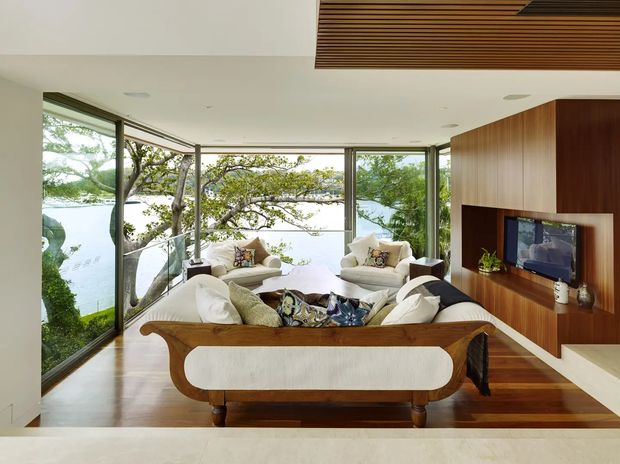 A bright, refined living room with extensive floor-to-ceiling glazing overlooking a leafy waterfront setting. The room features warm timber flooring, a large sculptural upholstered sofa, two cream lounge chairs, and a timber feature wall incorporating a recessed television and fireplace. A slatted timber ceiling detail adds architectural interest, while the open arrangement creates a strong connection to the landscape.