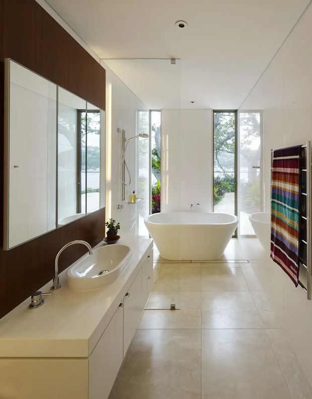 Bright contemporary bathroom with a freestanding oval bathtub positioned before tall windows, a long white vanity with countertop basin and mirrored wall cabinetry, a glass shower enclosure, and a colourful striped towel rack. Large pale stone-look floor tiles and white surfaces create an airy, refined interior, with dark timber wall panelling adding contrast.
