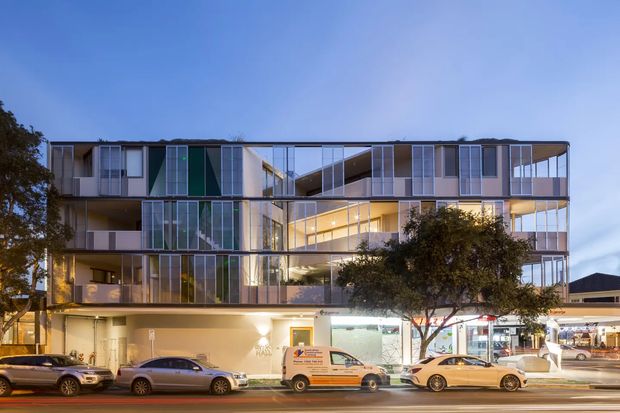 Street-level dusk view of a contemporary multi-storey mixed-use building with extensive floor-to-ceiling glazing, projecting balconies, transparent corner volumes, and illuminated ground-floor commercial tenancies. Vehicles and street activity occupy the foreground, while mature street trees partially screen the facade.
