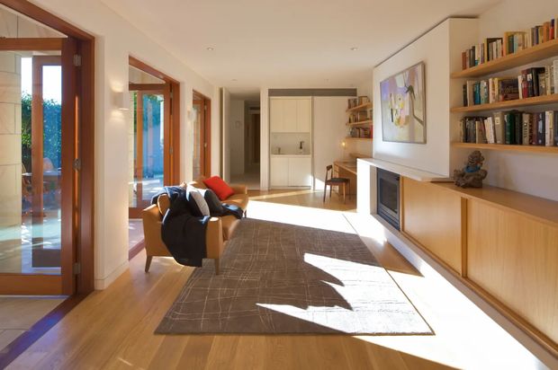 Bright, elongated living room with warm timber flooring, built-in shelving and cabinetry, a contemporary fireplace, armchairs, a large area rug, and abundant sunlight entering through timber-framed glazed doors. A hallway and adjacent spaces are visible in the background.