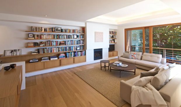 Warm, light-filled living room with extensive built-in timber shelving and cabinetry, a central fireplace, pale upholstered sofas, a large low coffee table, and broad timber-framed windows with horizontal blinds. The room has timber flooring, a neutral rug, and a recessed tray ceiling with concealed lighting.