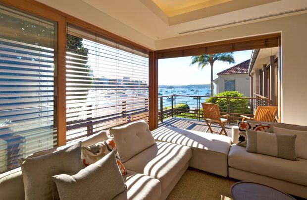 Sunlit living room with a large cream sectional sofa and lounge chairs opening onto a waterfront balcony. Timber-framed windows and doors with horizontal blinds surround the room, while a recessed tray ceiling adds architectural detail. The balcony has timber outdoor furniture and a black metal balustrade, with a bright harbour or coastal view beyond.