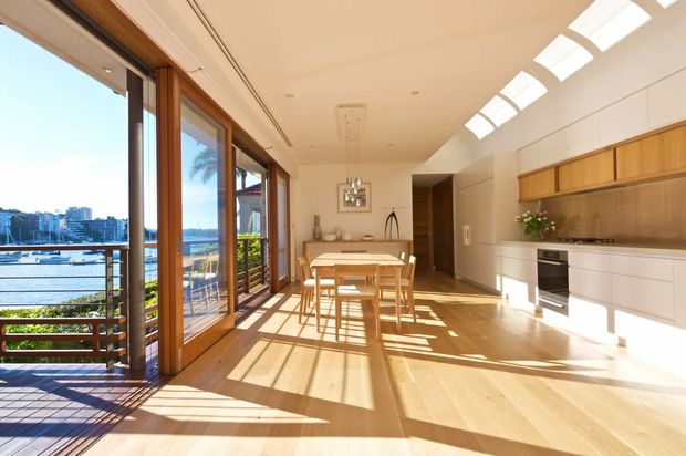Bright open-plan kitchen and dining area with extensive timber-framed glazing opening onto a balcony overlooking a marina or waterfront. The room features light timber flooring, white built-in cabinetry, a timber kitchen backsplash and upper joinery, a central dining table, and linear skylights or clerestory glazing that fill the space with sunlight.